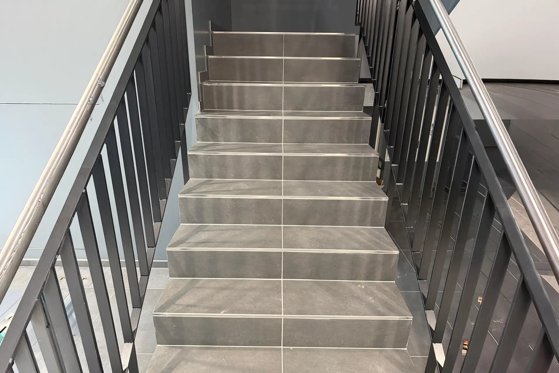 Modern geflieste Innentreppe mit großformatigen grauen Fliesen und schwarzem Metallgeländer.