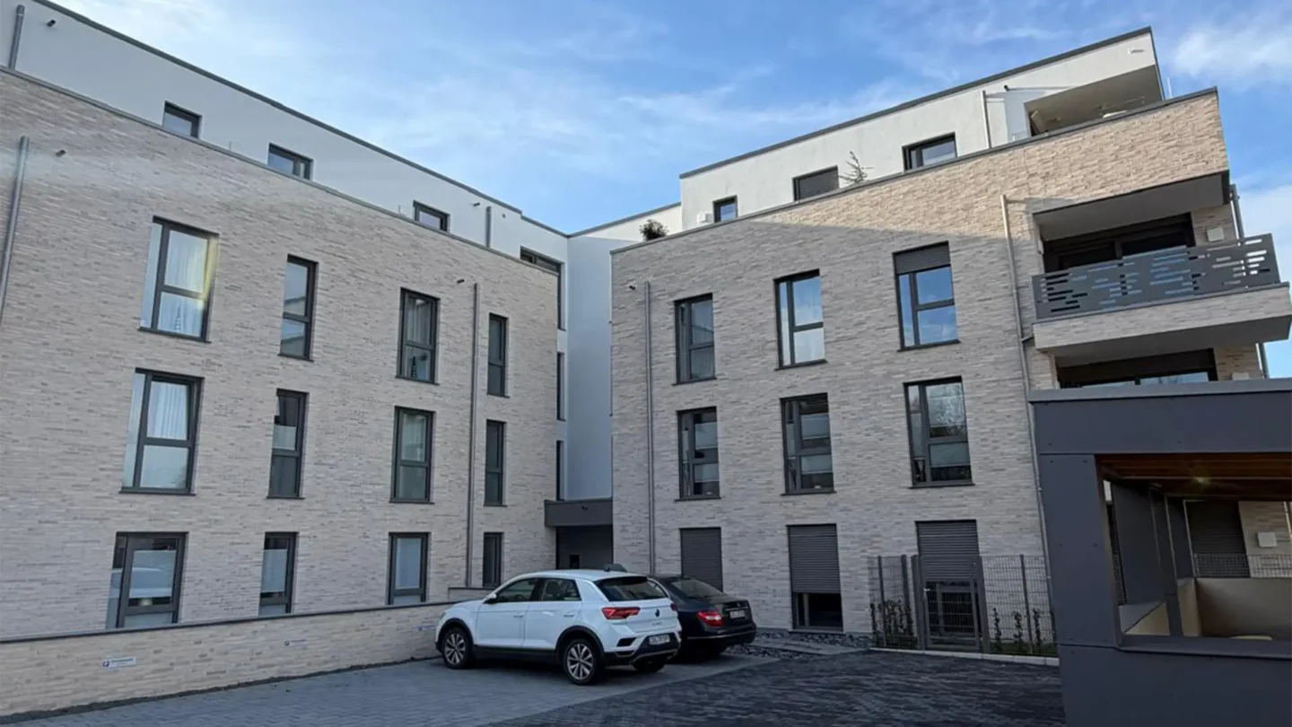Innenhof eines modernen Mehrfamilienhauses mit heller Klinkerfassade, großen Fenstern und Balkonen. Im gepflasterten Hof stehen zwei geparkte Autos, darüber spannt sich ein blauer Himmel mit leichten Wolken. Das Gebäude vermittelt einen hochwertigen und zeitgemäßen Wohn- und Gewerbebau.